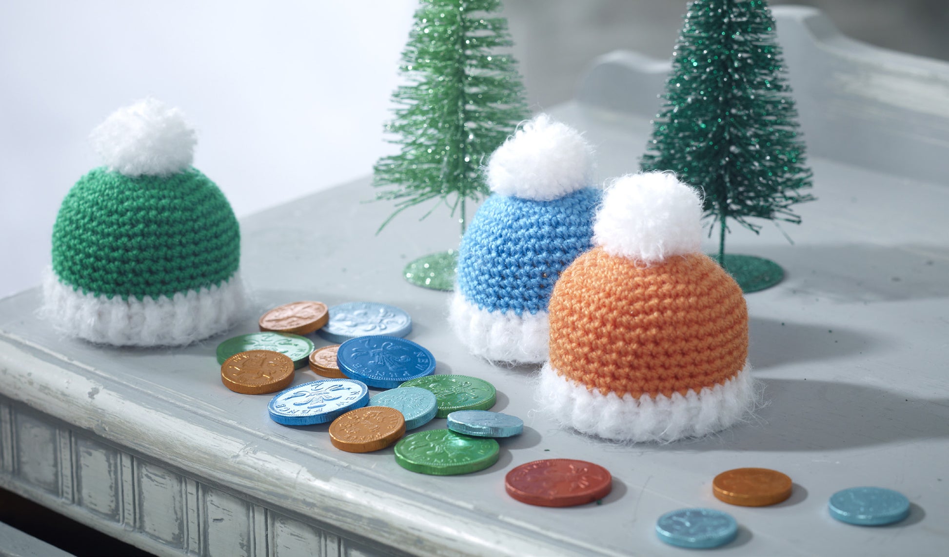 Bobble hat confectionary covers. Little hats perfects to top gifts. In this pattern there is one green, one blue and one orange hat, all with white bobbles and trim. Each hat is approximately 8cm high x 21cm around.